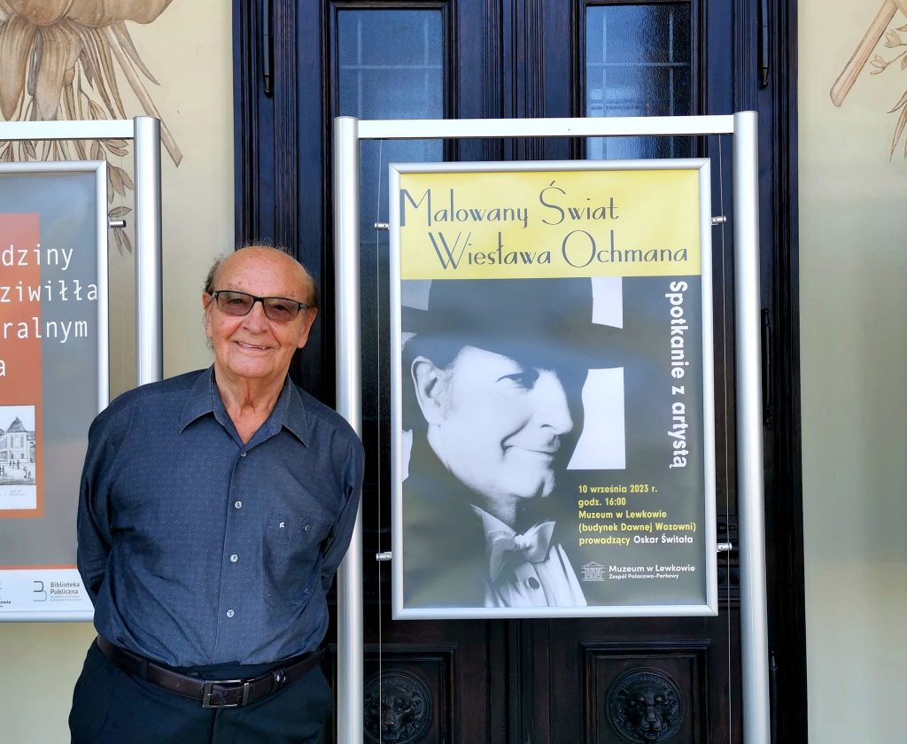 Wiesław Ochman w granatowej koszuli i granatowych spodniach stoi obok plakatu promującego spotkanie z artystą.
Plakat przedstawia czarno-białe zdjęcie elegancko ubranego, młodszego Wiesława. Na górze na żółtym tle czarny napis Malowany świat Wiesława Ochmana. Z boku obok zdjęcia pionowo biały napis spotkanie z artystą. Pod spodem poziomo żółty napis 10 września 2023 r. godz, 16:00 Muzeum w Lewkowie (budynek Dawnej Wozowni), prowadzący Oskar Świtała. Poniżej na czarnym tle białe logo muzeum