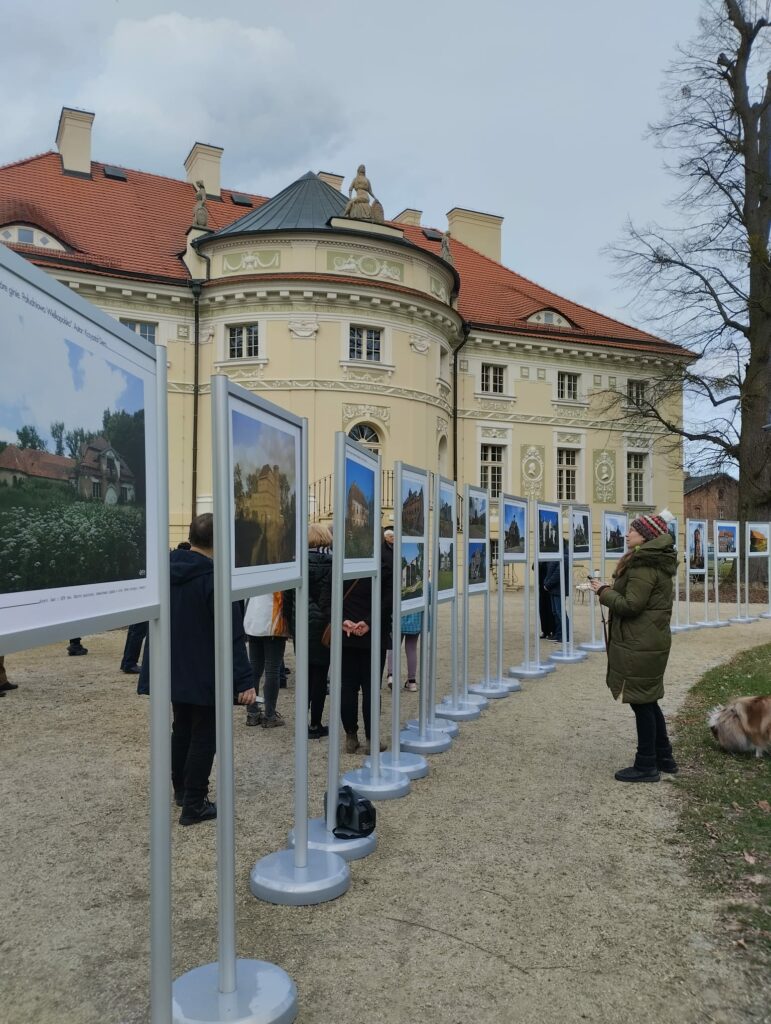 Zdjęcie przedstawia ludzi oglądających wystawę plenerową ustawioną na tyłach pałacu w lewkowie